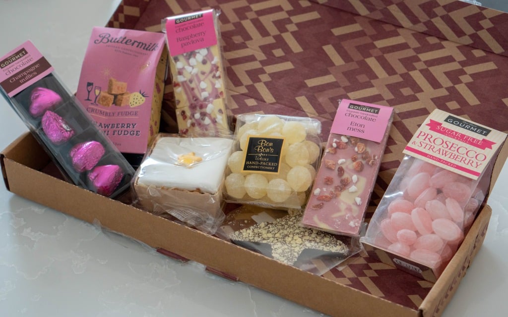 A selection of assorted sweets and chocolates in an open gift box, perfect for Mother’s Day, including pink heart-shaped chocolates, fudge, chocolate bars with toppings, boiled sweets, and a decorated iced biscuit.