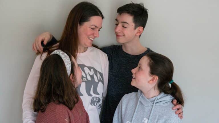 A woman and three children stand close together, smiling warmly at each other against a plain light-coloured wall, conveying affection and family bond—perfect for a 2020 Mother’s Day moment.