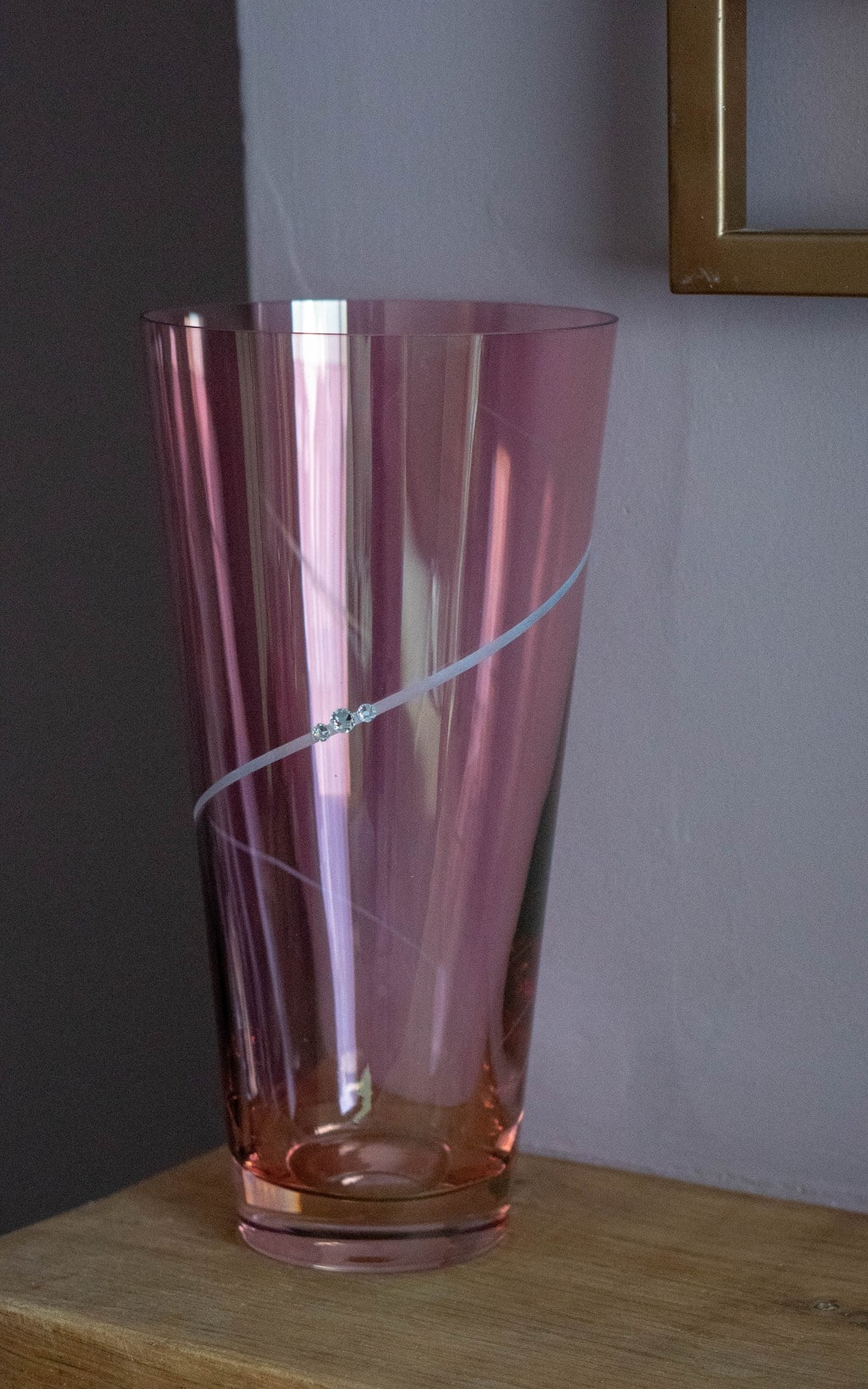 A tall, translucent pink glass vase with a thin, white diagonal stripe decorated with small sparkling gems sits on a wooden surface against a light purple wall—an elegant choice for any 2020 Mother’s Day Gift Guide.