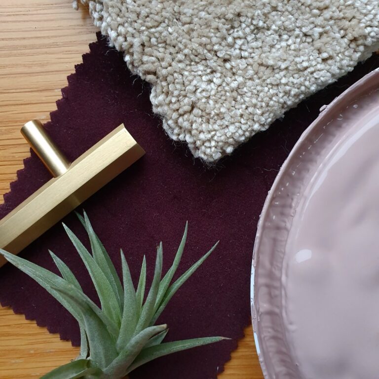 A beige carpet sample, brass drawer handle, green air plant, and mauve-pink paint sample sit on a wooden surface with a dark purple fabric square—perfect inspiration for your Pink and Gold Living Room.