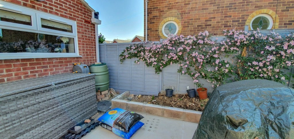 A small back garden perfect for garden planning, featuring a grey fence, pink flowering climber, potted plants, green water butt, wicker storage, a covered item, and a bag of compost. Brick houses rise in the background.