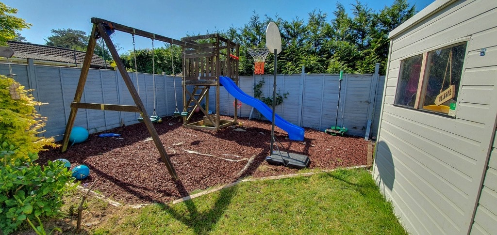 A back garden play area with a wooden swing set, slide, and climbing wall sits on bark chippings—perfect for fun or inspiring new garden ideas. Nearby, a white garden shed awaits your next gardening project, with green shrubs and trees lining the fence.
