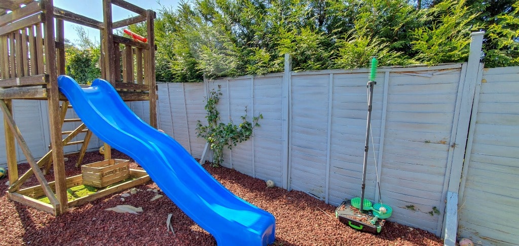 A playground with a blue slide attached to a wooden climbing frame sits on red gravel next to a white fence and green bushes—perfect inspiration for creative garden planning. Two green-handled pogo sticks lean against the fence.