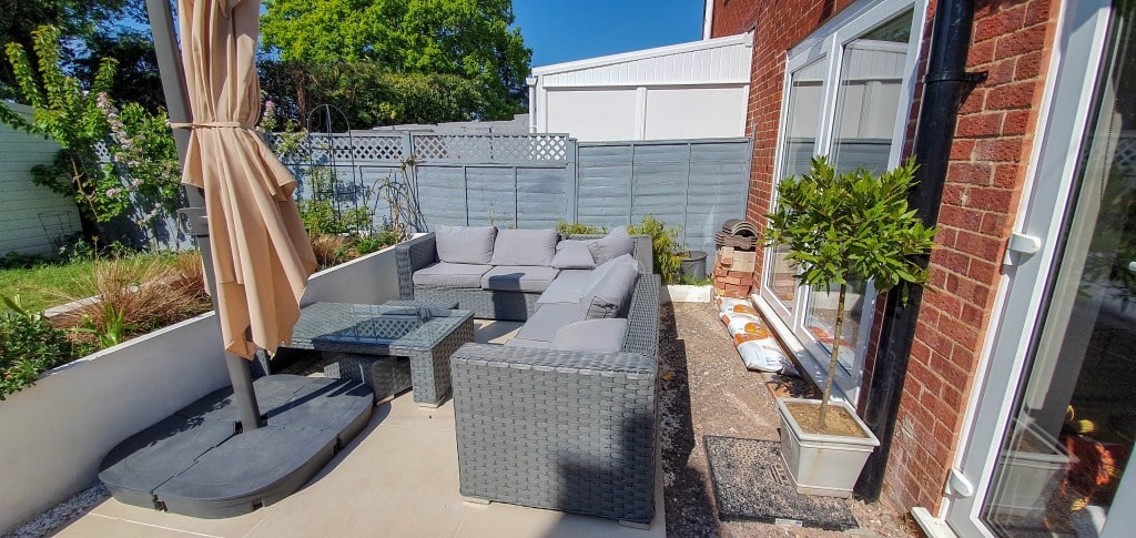 A patio with grey wicker furniture, including a sectional sofa and armchair, surrounds a glass coffee table. A parasol stands nearby, while plants and garden supplies hint at ongoing gardening against a fenced backdrop.