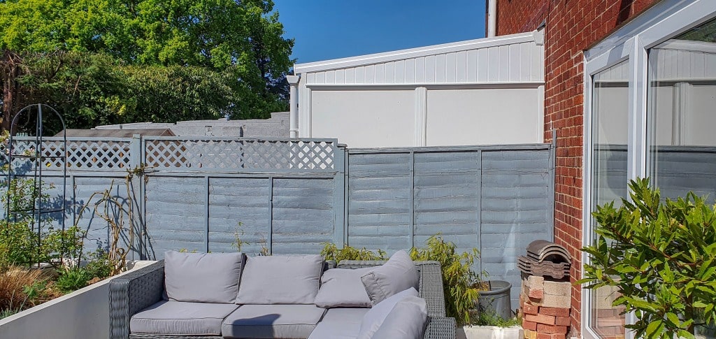 A sunny back garden patio with a grey outdoor sofa set against a tall grey fence, green plants nearby, and a stack of bricks beside the sofa—perfect for relaxing or working on your latest garden planning ideas. A red brick house and blue sky complete the scene.