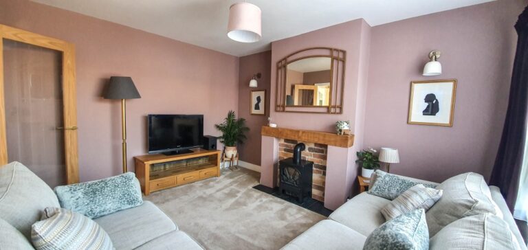 A cosy pink living room with beige sofas, a TV on a wooden stand, and a brick fireplace with a mirror above. Framed silhouette art decorates the soft pink wall, whilst lamps, plants, and natural light complete this warm Living Room Reveal.