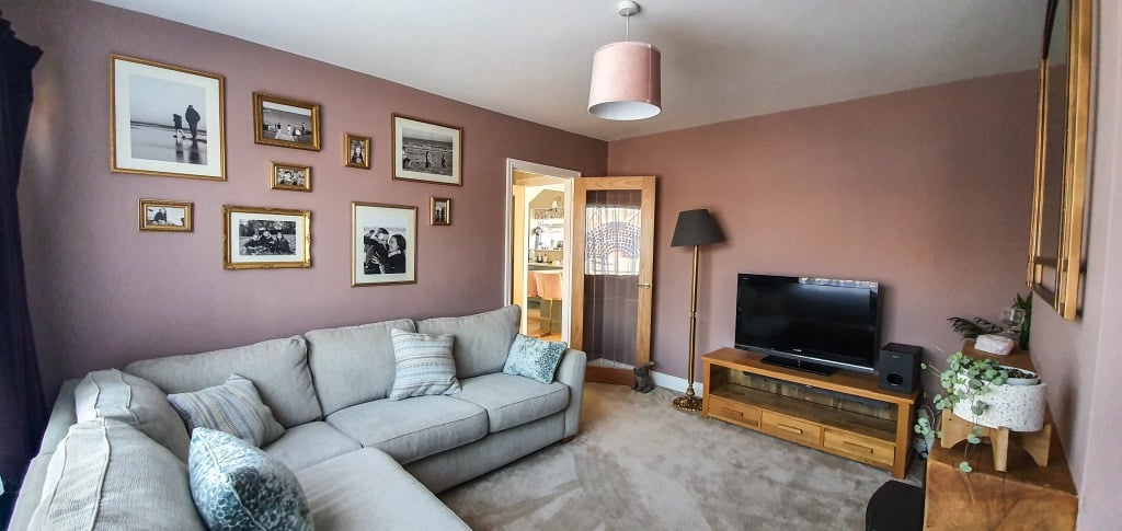 A cosy gold living room with a light grey sectional sofa, patterned cushions, framed photos on a mauve wall, a wooden TV stand with TV, floor lamp, and a doorway leading to another room creates the perfect living room reveal.