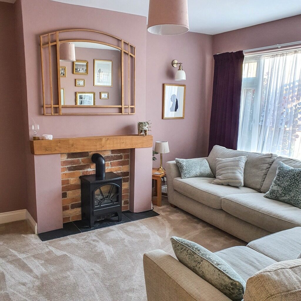 Cosy Pink Living Room reveal with beige sofas, a wooden mantel fireplace featuring a black stove, wall art, a large mirror, and light brown carpet. Pinkish walls, sheer curtains, and a stylish side table with lamp complete the inviting space.