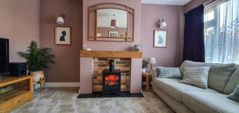 A cosy living room reveal with a small electric fireplace beneath a wooden mantelpiece, a large mirror above it, two silhouette portraits on either side, a grey sofa, potted plant, and light streaming through sheer curtains.
