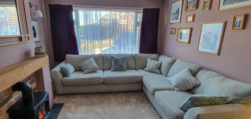 A cosy pink living room with a light beige U-shaped sofa, several cushions, framed pictures on mauve walls, a fireplace with a wood-burning stove, and large windows with sheer and dark curtains letting in daylight.