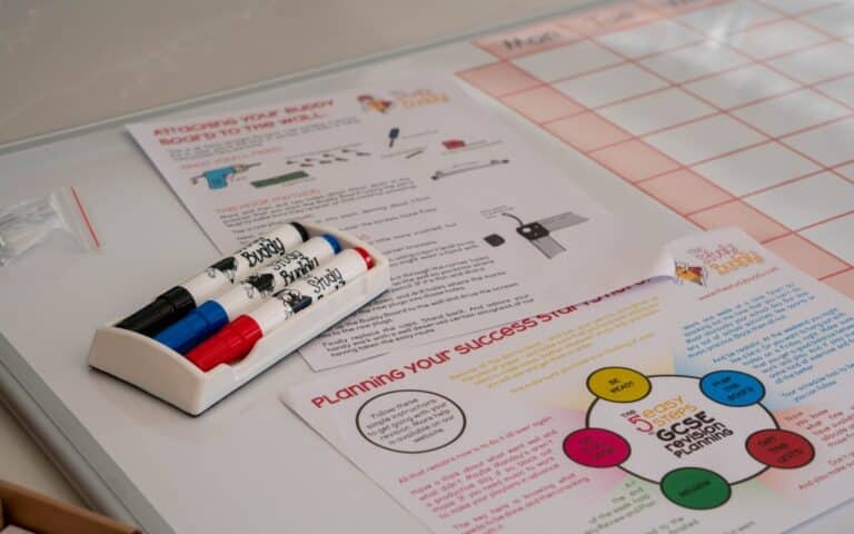 A Study Buddy Board whiteboard with a grid chart, colourful markers in a holder, and printed planning sheets with diagrams and text about success strategies helps you stay focused during lockdown at your desk.