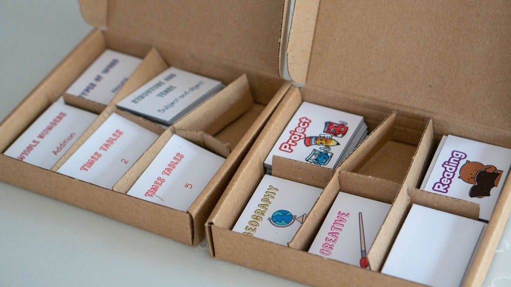 An open cardboard box with dividers holds stacks of labelled cards, including PROJECT, GEOGRAPHY, READING, and other subjects—like a Study Buddy Board to help you stay focused—with some featuring colourful illustrations.