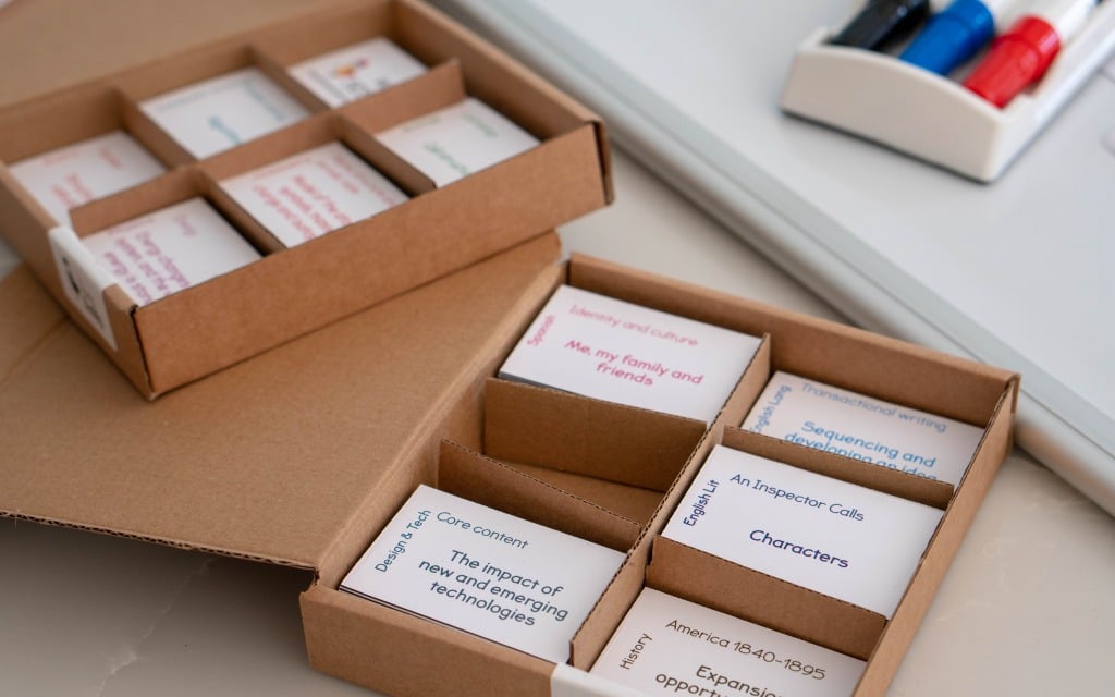 A cardboard box is divided into compartments, each containing stacks of small cards with educational topics and phrases printed on them. The Study Buddy Board and marker tray are visible in the background, helping students stay focussed during lockdown.
