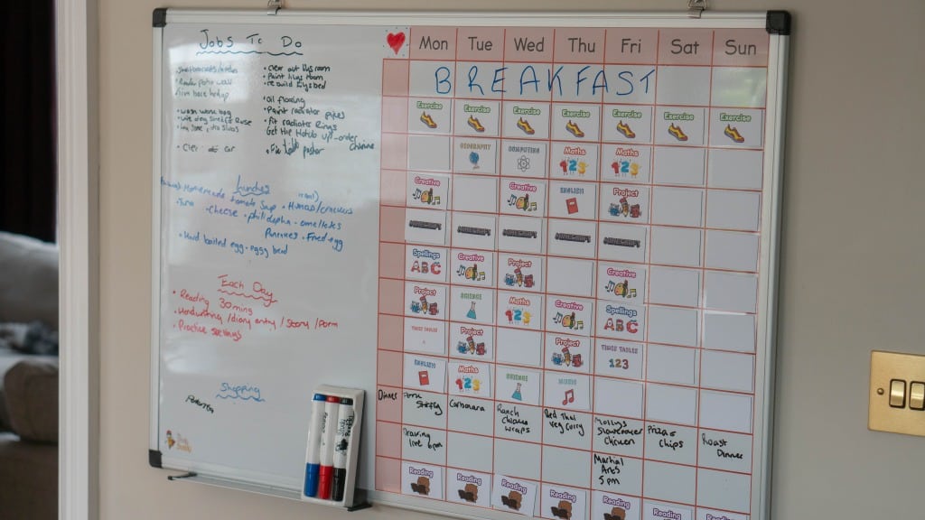 A Study Buddy Board with a “Jobs To Do” list and a colourful weekly breakfast schedule shows days, meals, and cute images; markers stay attached to the board, which is mounted on the wall near a light switch to help you stay focused during lockdown.