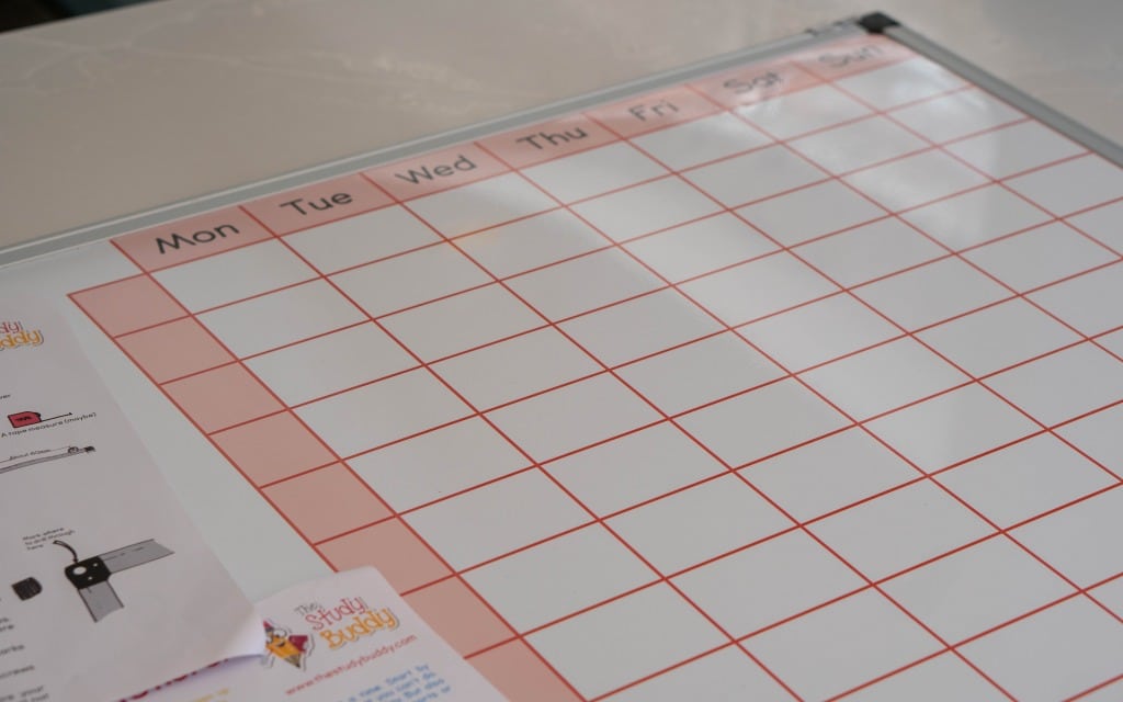 A blank Study Buddy Board whiteboard calendar with red grid lines and columns labelled Monday to Sunday. Papers, including an instruction sheet, are partially visible on the left side—perfect to help you stay focused, even during lockdown.