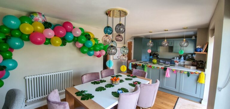 A dining room decorated for an 11th Birthday party with colourful balloons, paper leaf placemats, and tassels. The table is set for six, and the kitchen in the background also has festive Happy Birthday decorations.