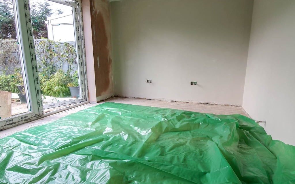 A room undergoing renovation with bare walls and floor, a large window to the left, and a green plastic sheet covering part of the floor—an ideal space for DIY wood flooring projects. Lush plants are visible outside through the window.
