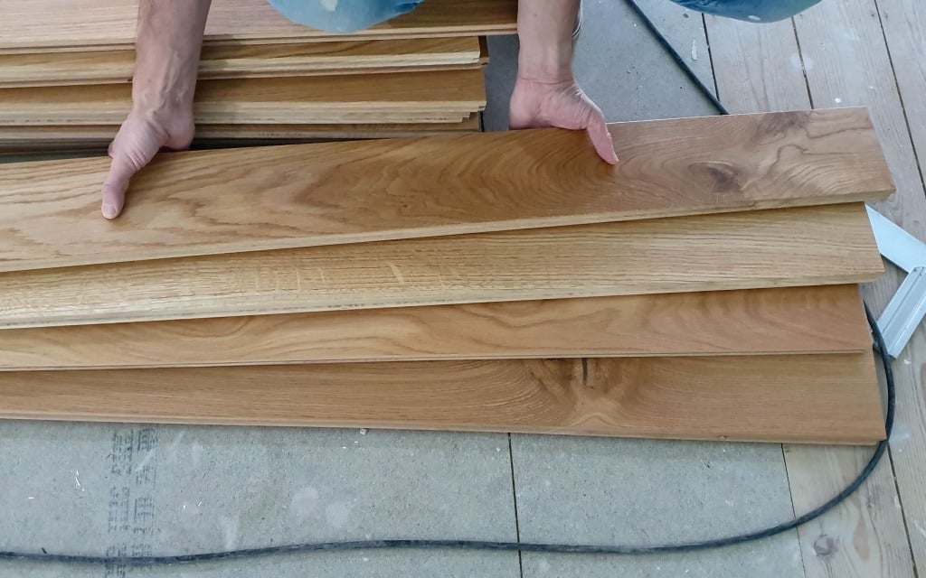 A person’s hands hold and organise several solid wood flooring boards, stacked neatly on a floor under construction.