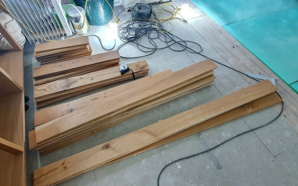 Stacks of wooden floorboards, an electric saw, power leads, and a green underlay are spread out on a partially installed solid wood flooring project in a room undergoing renovation.