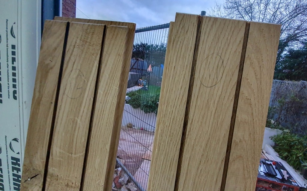 Two wooden planks with three parallel grooves each stand upright outdoors near a construction site, ideal for laying flooring. Pencil markings are visible on the planks, with a garden, fence, and tools in the background.