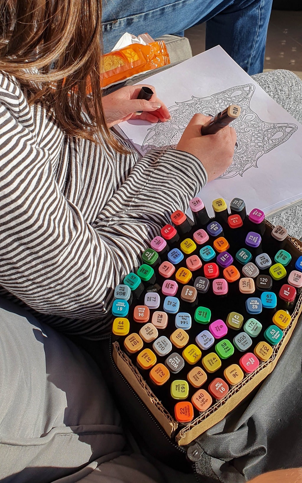 A person wearing a striped shirt colours a detailed mandala in a colouring book, using a large set of markers on their lap—sunlight streaming over their hands. It's a perfect relaxing activity for learn from home or home-schooling settings.
