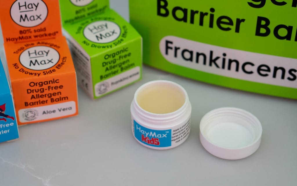 A small open pot of HayMax Kids balm sits on a surface, surrounded by colourful HayMax product boxes—perfect for children who learn from home—with part of a green sign that reads “Barrier Balm” and “Frankincense.”.