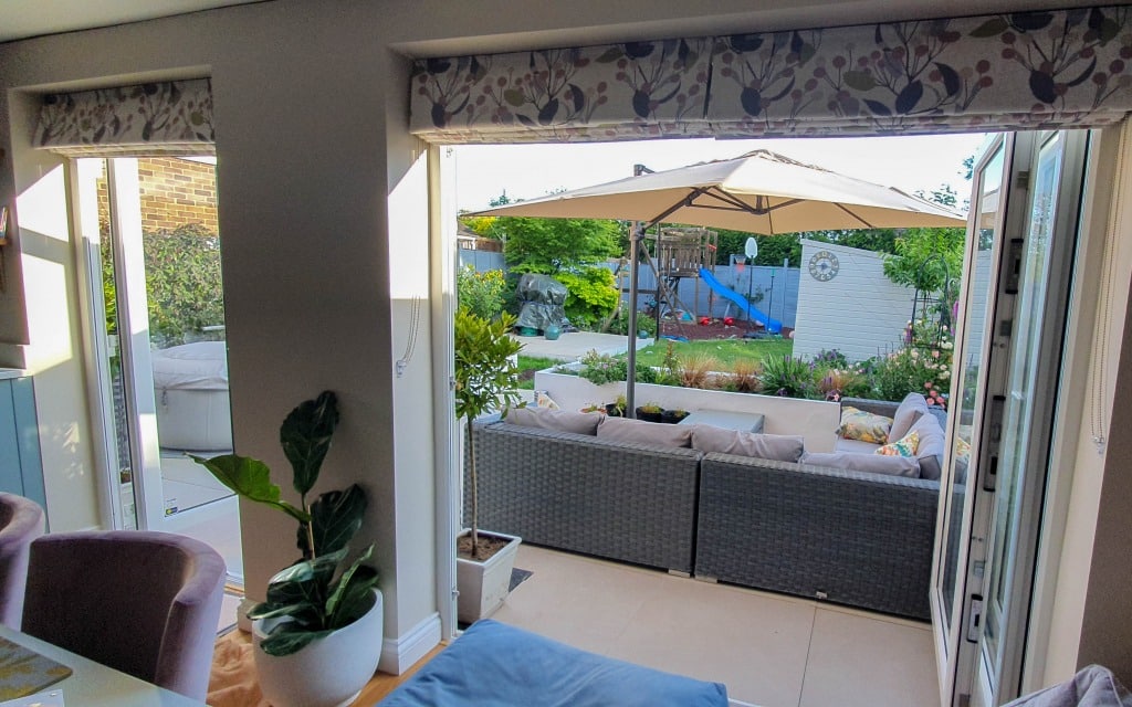 A bright, modern living room opens to a patio with grey outdoor sofas and a large parasol. Beyond the patio is my garden in May, filled with green plants, flowers, and a children’s play area perfect for enjoying Garden This Month.