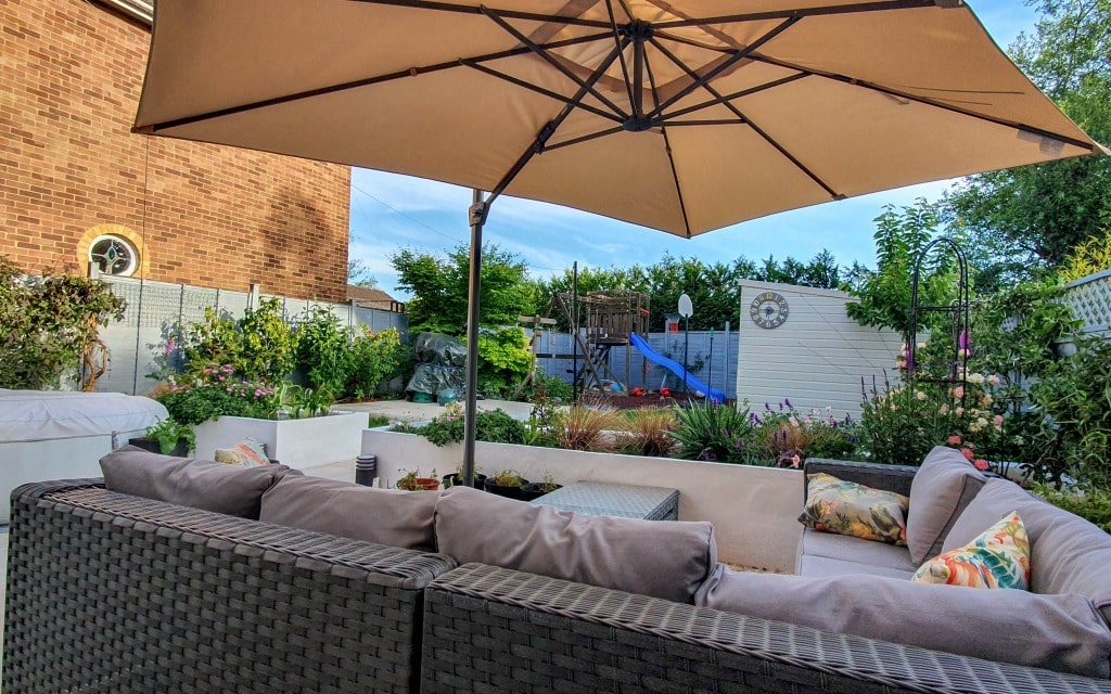 A cosy back garden patio in My Garden features a large parasol, grey outdoor corner sofa, decorative cushions, a table, and lush landscaping. In the background, a climbing frame with a slide and fence adds charm—perfect for relaxing this May.