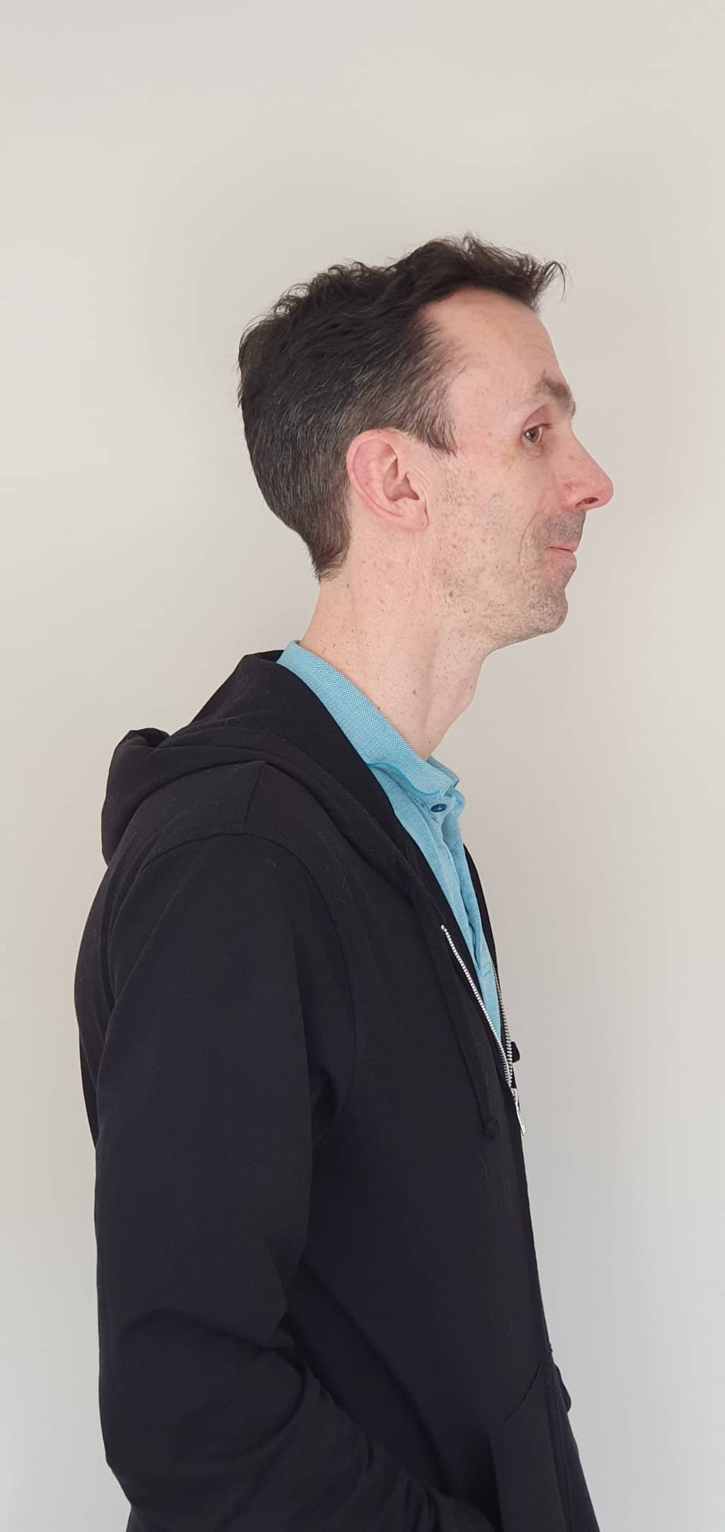 A man with short dark hair wearing a black hoodie over a blue collared shirt stands in profile against a plain light-coloured background, perfect for creating striking Personalised Silhouette Prints.
