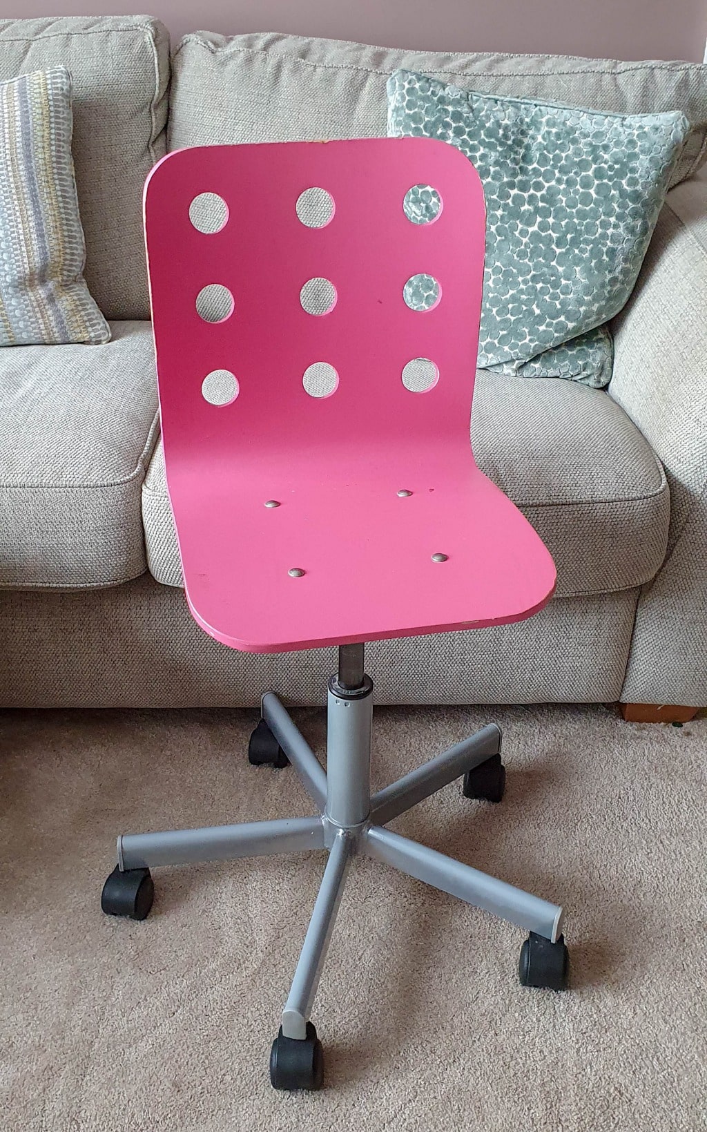 A pink swivel chair with circular cut-outs in the backrest and a silver base on castors, perfect for a small bedroom, is positioned in front of a beige sofa with patterned cushions on a carpeted floor.