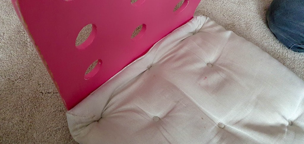 A pink IKEA Jules chair with circular cut-outs on the backrest and a white, tufted cushion seat sits on a light-coloured carpet. Part of a person's knee in jeans appears in the top right corner, suggesting it's being used as a desk chair.