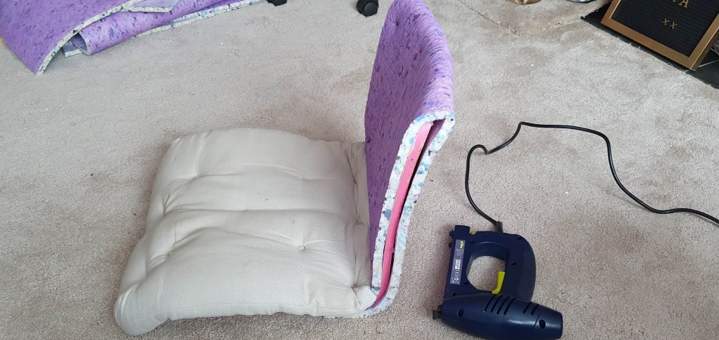 A cushioned floor chair with a beige seat and purple backrest—similar to an upholstered IKEA chair project—sits on a carpet next to a blue electric stapler. An amp and more cushions are visible in the background.