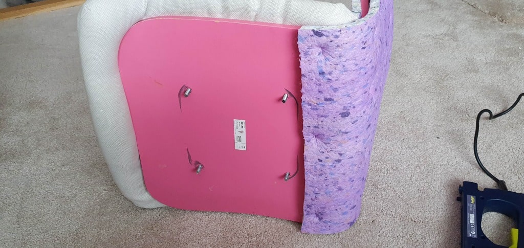 A chair seat, likely from an IKEA Jules desk chair, is flipped upside down on a carpeted floor. The pink bottom panel is visible, with upholstery partially removed and purple foam padding exposed. A staple gun sits to the right.