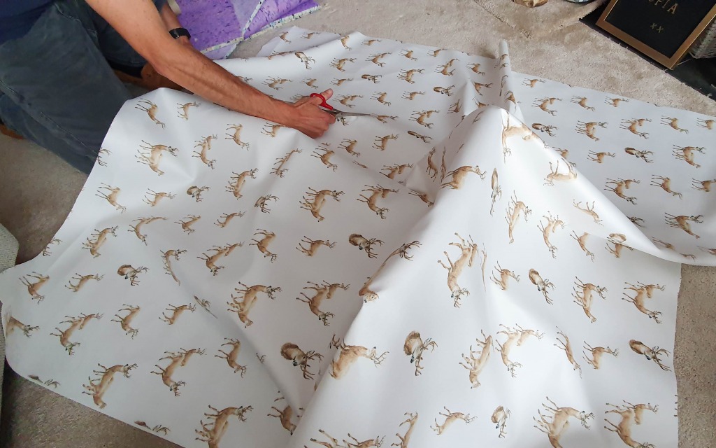 A person is cutting a large sheet of white paper with brown reindeer prints on a beige carpet, using red-handled scissors—perhaps preparing to upholster an IKEA Jules desk chair for a festive DIY project.