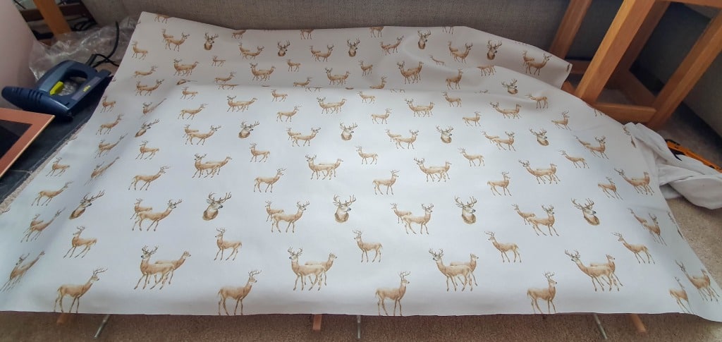 A large piece of fabric with a repeating pattern of illustrated deer, laid out on a flat surface with furniture and tools visible around it, ready for upholstering the headboard of an IKEA MALM bed.