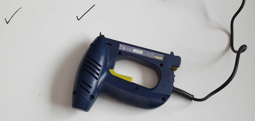 A blue electric staple gun with a yellow trigger is lying on a white surface, ideal for upholstering projects like creating a custom headboard for an IKEA MALM bed. Its black power lead extends to the right, near two black tick marks.