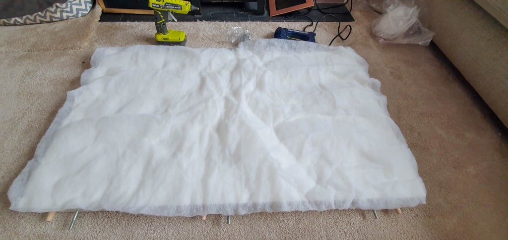 A large piece of white wadding is spread out on a tan carpeted floor, with tools like a green power drill and screws nearby—likely for upholstering a bed headboard, perhaps for an IKEA MALM bed DIY project.