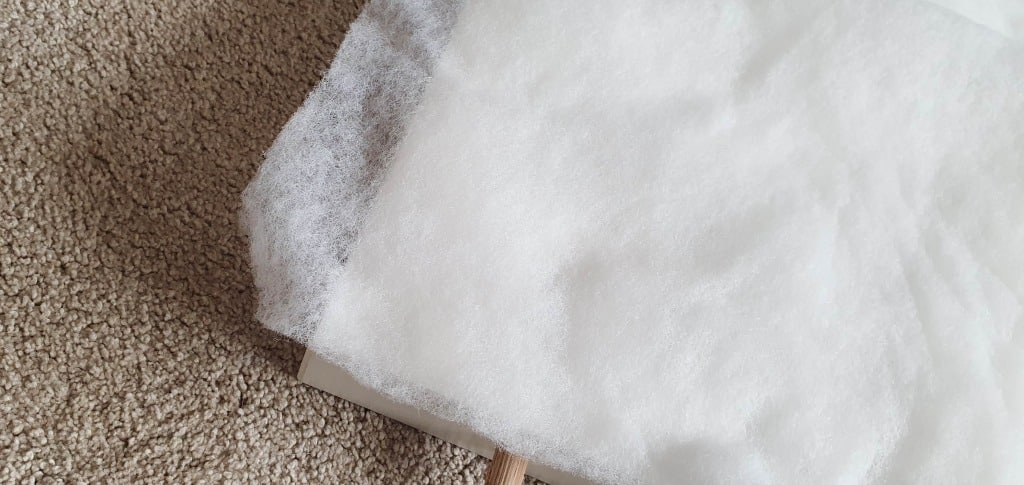 A piece of fluffy white wadding or fibre filling lies partially folded on a beige carpeted floor, with part of it hanging over the edge of an IKEA MALM bed headboard, ready for upholstering.