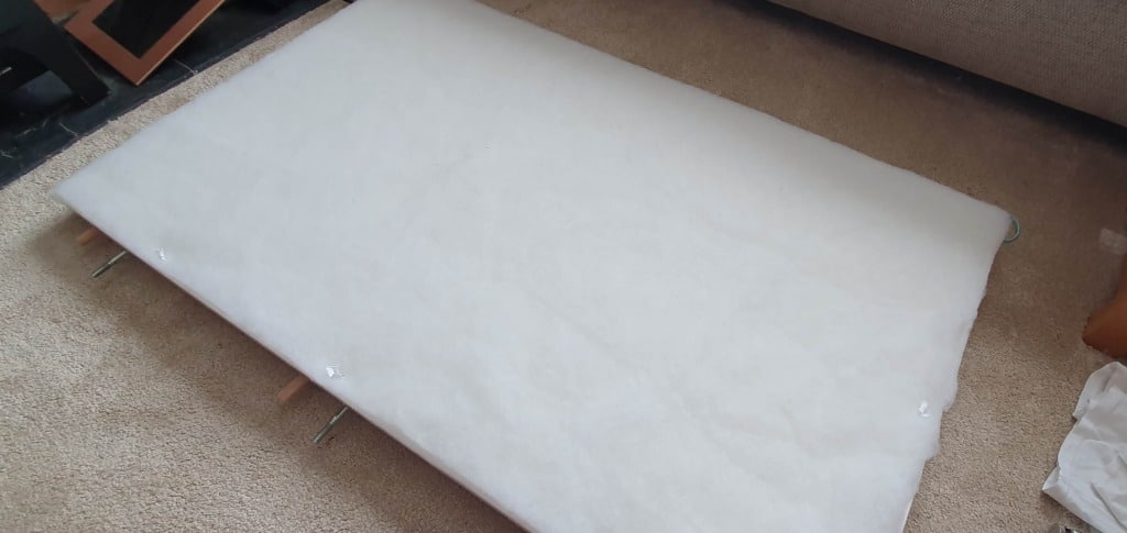 A large rectangular board, possibly from an IKEA MALM bed headboard, lies on a beige carpeted floor, covered with white wadding as if being prepared for upholstering headboard DIY. Metal bolts and wooden supports can be seen at the edges.