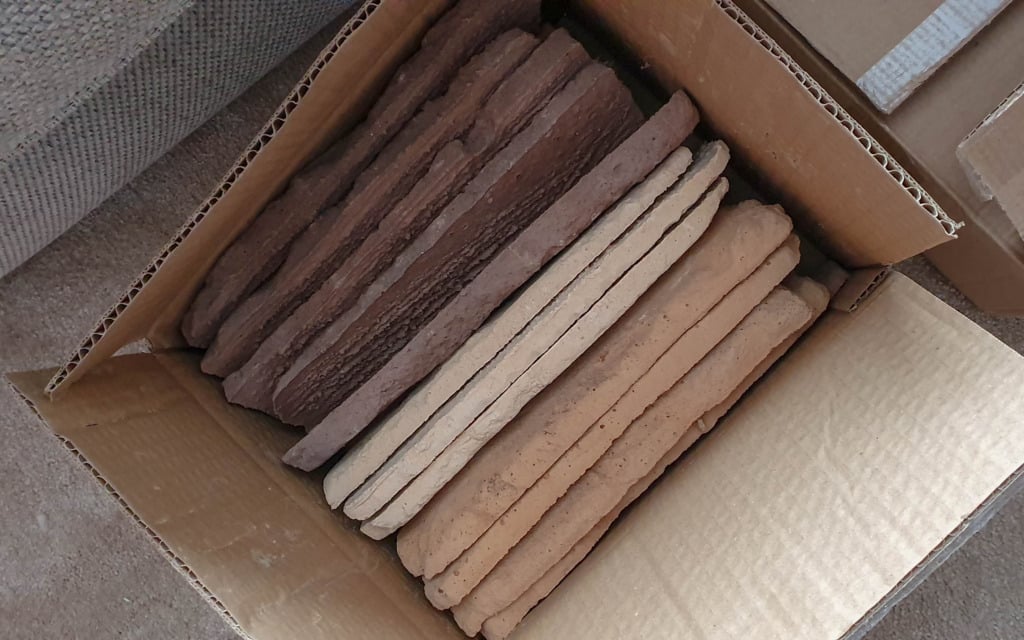 Cardboard box containing stacked, rectangular clay tiles in two colours: a few dark brown and more light tan tiles, perfect for a Kuci Design brick slip fireplace. The box is sitting on a carpeted floor.