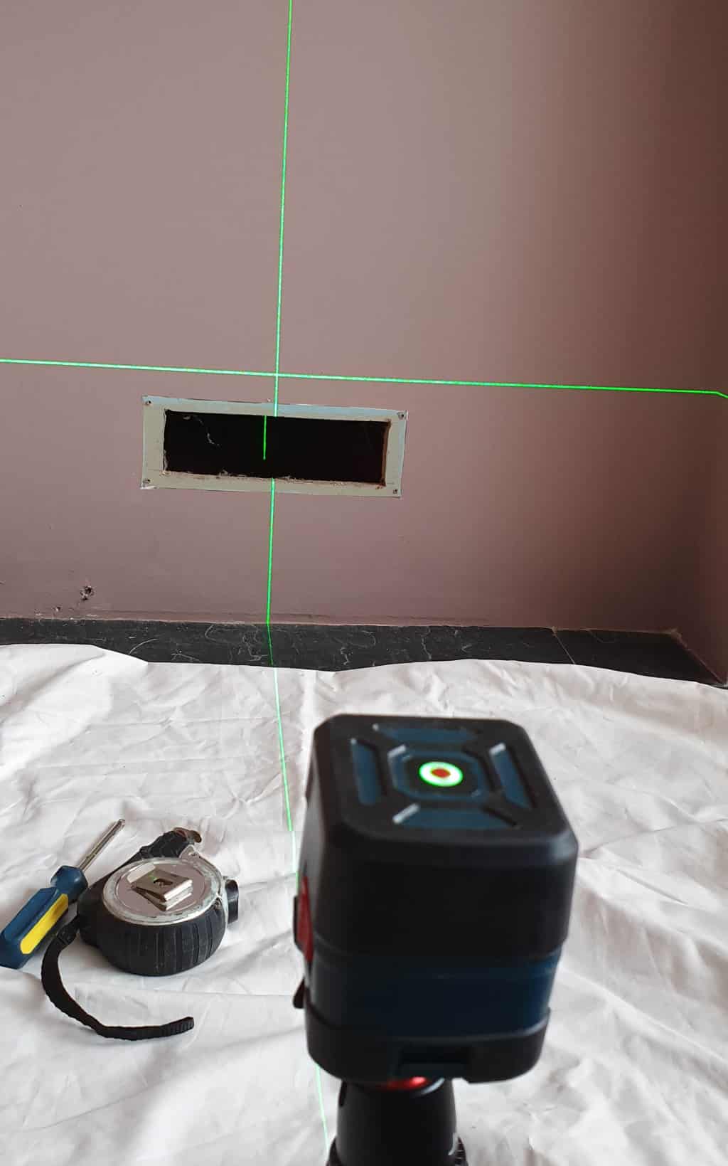 A green laser level projects horizontal and vertical lines on a wall with a rectangular hole, ideal for aligning brick slips. A screwdriver and measuring tape lie on a white sheet on the floor, reflecting Kuci Design’s precision approach.