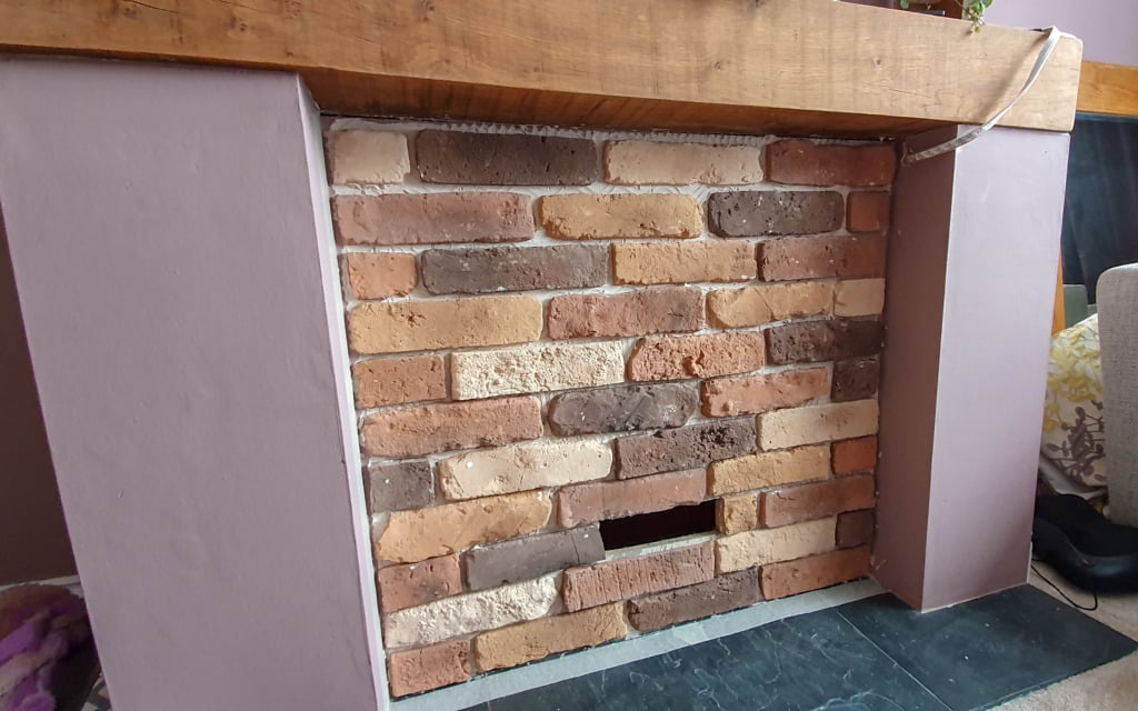 A fireplace filled with brick slips in various shades of brown, beige, and red, with one horizontal brick missing near the bottom centre. A wooden mantelpiece by Kuci Design rests above this unique brick slip fireplace.