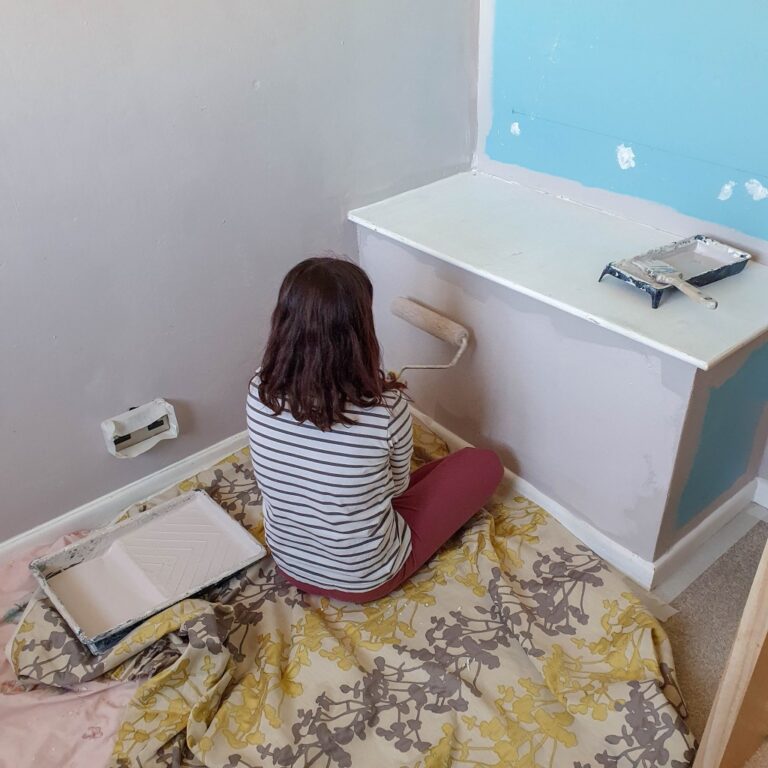 A person with shoulder-length hair, wearing a striped shirt and red trousers, sits on a patterned sheet while painting a wall and cupboard for a Small Bedroom. Paint trays and tools are scattered nearby.