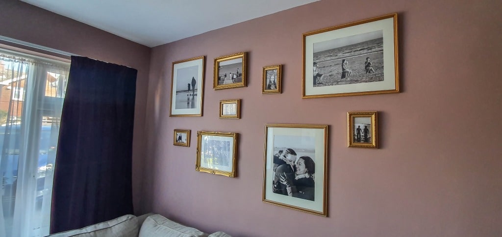 A pink wall displays several black and white photographs in gold frames, arranged gallery style above a beige sofa, next to a window with sheer and dark curtains.