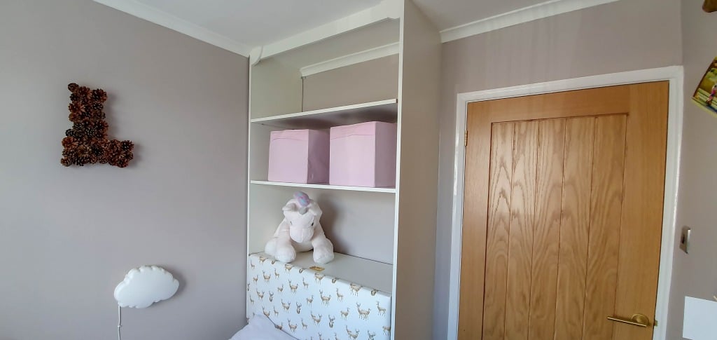 A light grey small bedroom corner with a wooden door, a white shelf holding two pink storage boxes, a plush unicorn toy, and a cloud wall light above giraffe print bedding. A decorative L hangs on the wall in this cosy Bedroom Reveal.