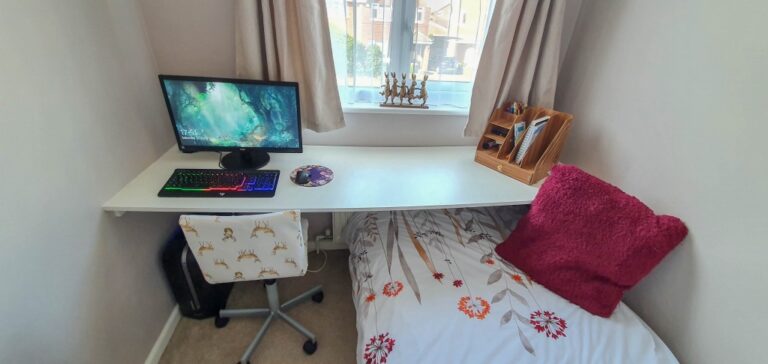 A tidy small bedroom workspace with a monitor, keyboard, and mouse on a white desk sits next to a single bed with floral bedding and a pink pillow. A window with curtains and a wooden organiser are above the bed.