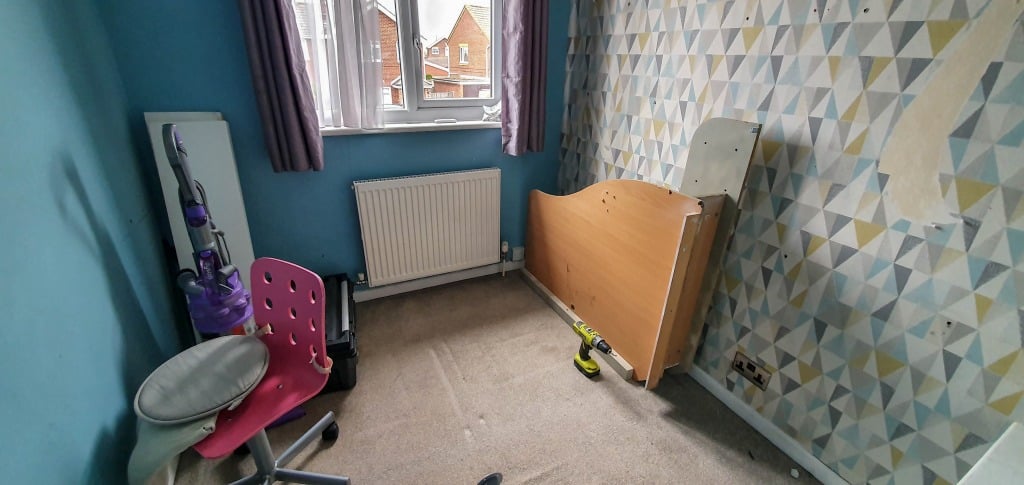 A Small Bedroom with blue geometric-patterned walls, purple curtains on the window, a pink chair, vacuum cleaner, partial bed frame, and tools scattered on the carpet awaits your Bedroom Reveal.