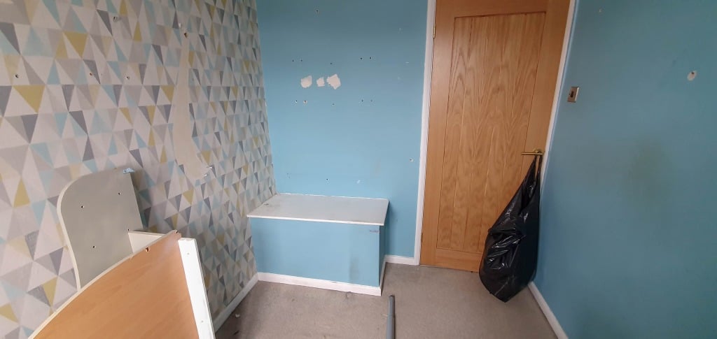 A small bedroom with blue walls, patterned wallpaper on one wall, a wooden door with a black bin bag on the handle, and some furniture against the walls—perfect for a cosy Bedroom Reveal.
