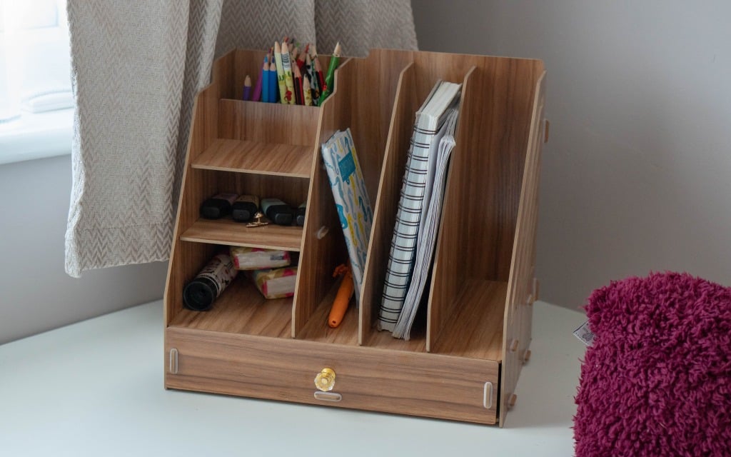A wooden desk organiser holds coloured pencils, pens, notebooks, and small toys on a white desk near a window with curtains and a purple cushion—perfect for your next Small Bedroom or Bedroom Reveal.