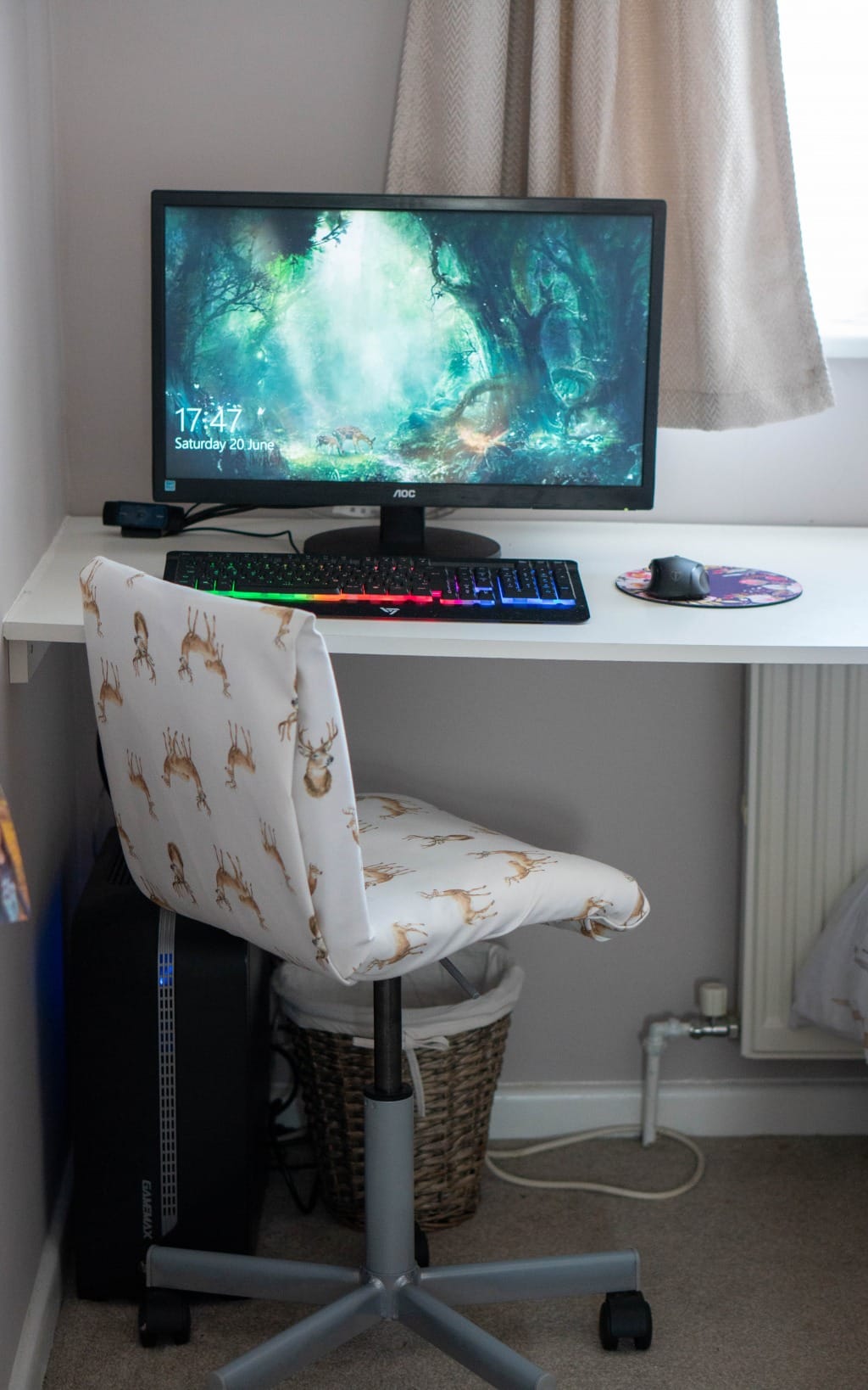A computer desk with a monitor displaying a forest scene, a colourful backlit keyboard, and a mouse on a printed mouse mat. Perfect for a small bedroom, the setup includes a white chair with a deer pattern and a basket underneath.